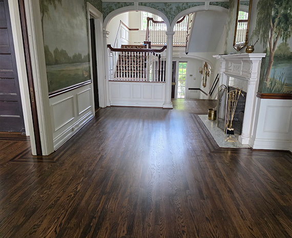 Elegant foyer with dark hardwood floor, white paneled walls, arched doorways, and a staircase railing visible in the back, plus a decorative fireplace on the right.
