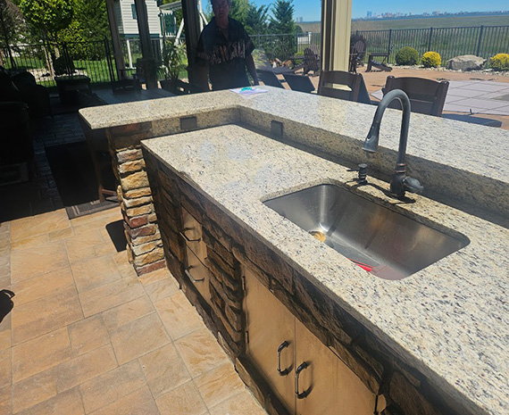 Outdoor kitchen island with granite countertop, stone base cabinets, and a stainless steel sink with a gooseneck faucet; patio and yard in the background.