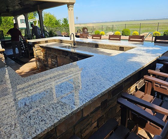 Outdoor stone-and-granite outdoor kitchen with a long bar, stools, and a view of a fenced field in the distance.