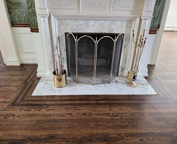 Marble fireplace with white mantle and brass-andiron fire screen; brass tool stand on the right and a brass bucket with tools on the left, atop dark hardwood flooring.