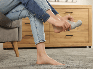 Person sitting on a sofa, putting on a gray sock over one bare foot in a living room setting.