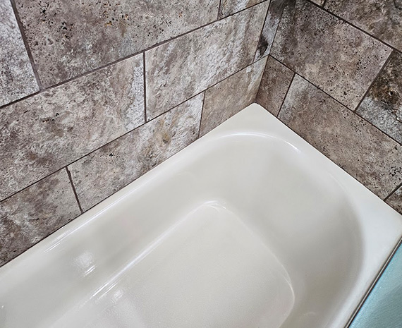 White enamel bathtub in a tiled bathroom corner with brown-gray stone tiles on the walls, seen from above edge view.