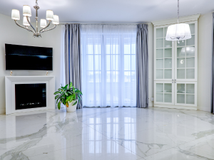 Modern living room with a white marble floor, fireplace, and wall-mounted TV, plus a potted plant near a large window with sheer curtains.